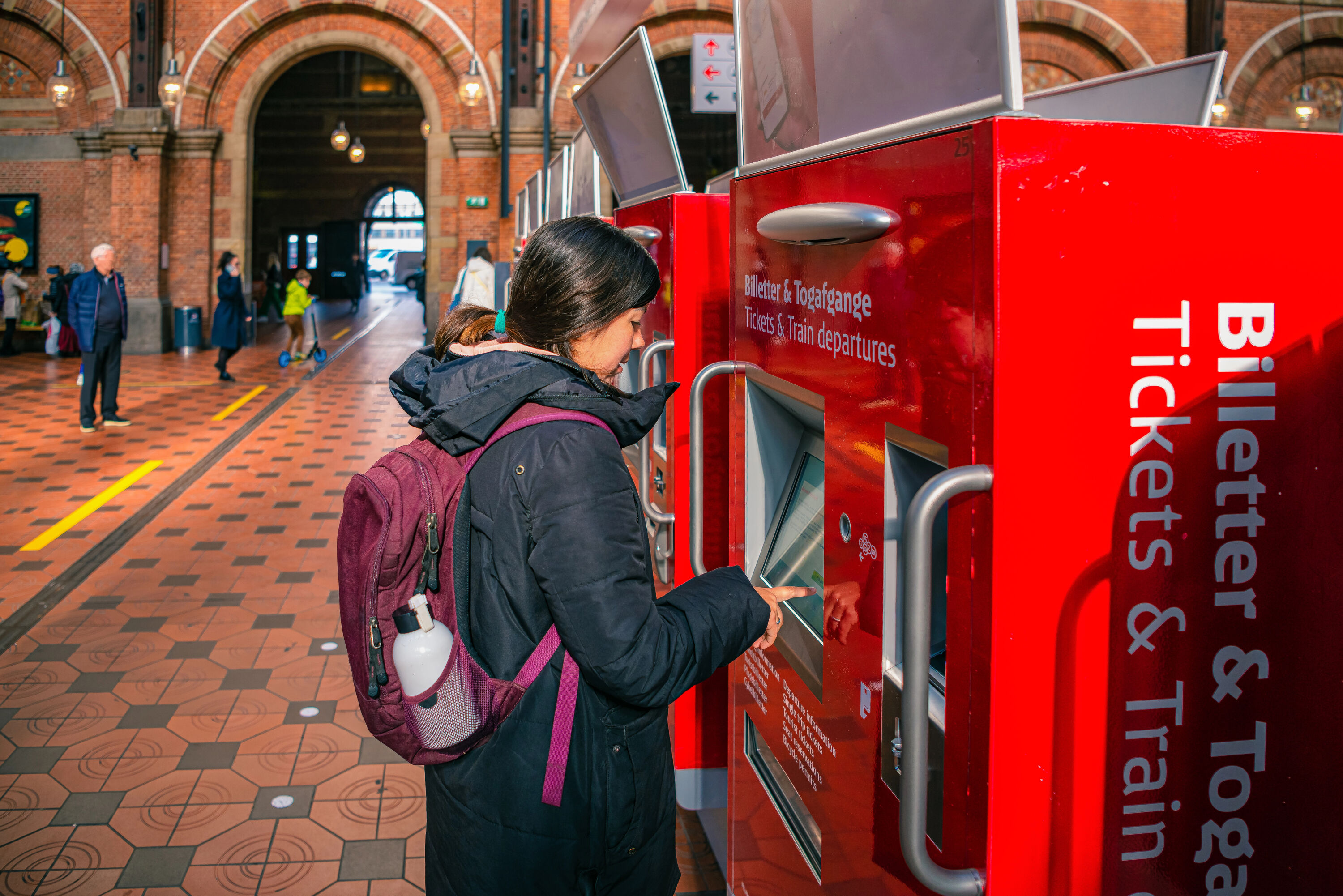 Young woman tourist buys a ticket for the metro, bus or train in a red vending machine in the main hall of the Copenhagen Central railway Station. Denmark